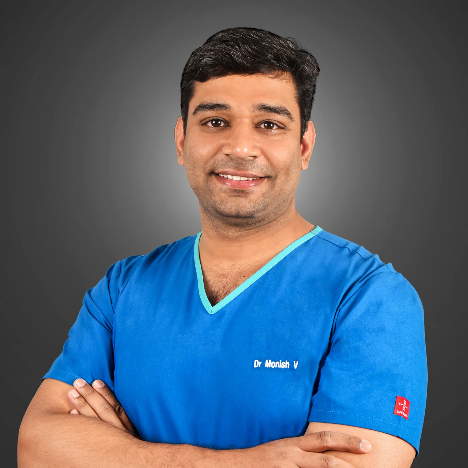 homepage 17 Dr. Monish V. posing in a blue medical scrub with a professional smile.